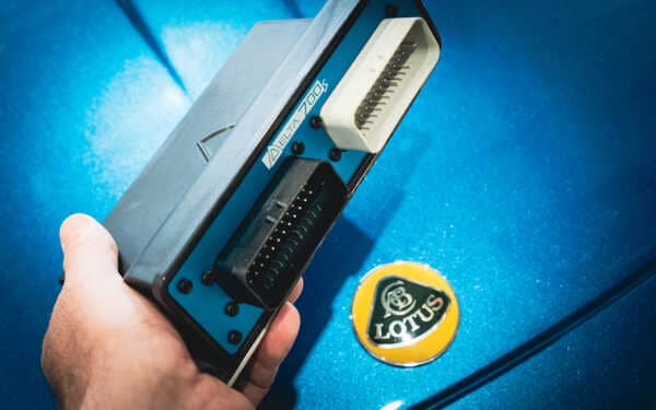 SCS Delta 700s ECU about to be fitted to a Lotus Elise S2. close up photo of the ECU being held in a man's hand against a beautiful metalic blue Lotus with the yellow Lotus badge below.