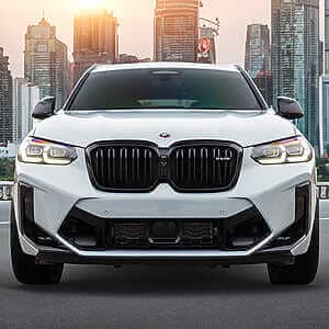 BMW F98 X4M Tuning on the S58 platform. Front view of white BMW SUV parked behind a city with skyscrapers backlit with sunset