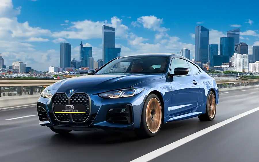 BMW M440i G22 Tuning Box B58TU Strat-2 2 BMW M440i G22 side photo of the car speeding down a city street. The car is dark blue with gold wheels, driving on a grey road with blue sky and tower blocks in the background