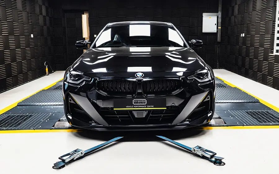 BMW M240i G42 Tuning Box B58TU Strat-2 1 BMW M240i G42 Tuning this is a front view of a black BMW in a grey sound proof cell. It is strapped down by two blue straps ready to be tested for power