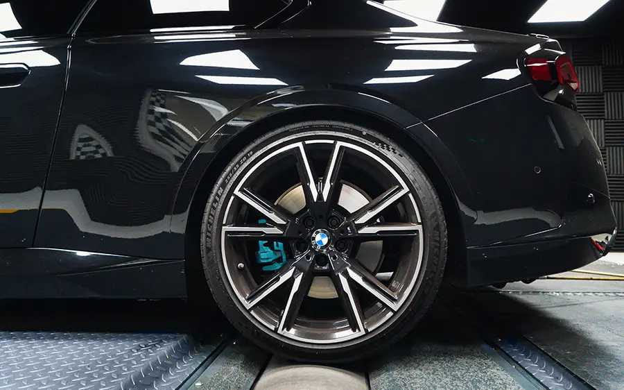 BMW M240i G42 Tuning Box B58TU Strat-2 4 Close up shot of the rear wheel of a car. The wheel is black and silver with a light blue brake caliper. The car is black